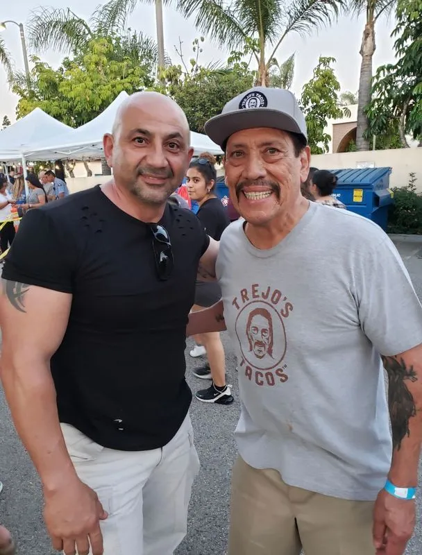 Two men are posing for a picture and one is wearing a shirt that says trejos tacos