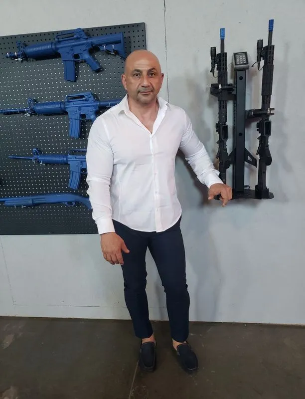Man in white shirt and dark pants stands by blue training rifles and a weapon rack.